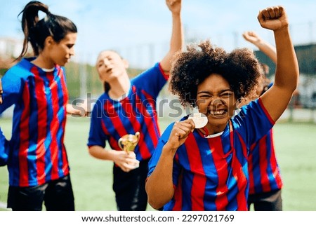 Cheerful African American soccer player with a gold medal at the stadium looking at camera. Her teammates are in the background. 