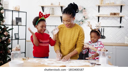 Cheerful African American woman in apron with little kids standing at table in home Christmassy decorated kitchen and having fun while making xmas cookies. Holidays concept