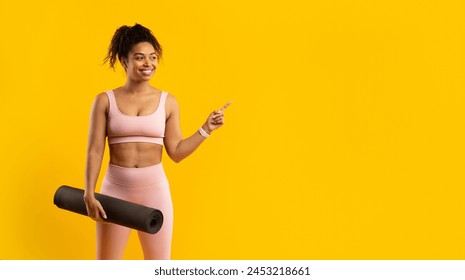 A cheerful african american woman in athletic wear holding a yoga mat and pointing to her side against a yellow background - Powered by Shutterstock