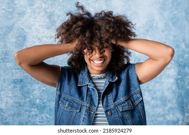 cheerful african american woman in denim vest touching curly hair on blue background - Powered by Shutterstock