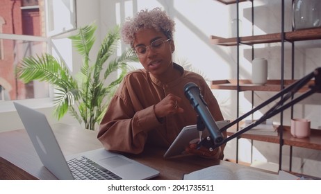 Cheerful african american woman podcaster records her voice into a microphone. Beautiful female radio host hosting a streaming podcast using a microphone and laptop in her home studio.
