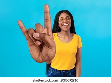 Cheerful African American Woman Showing Big Rock Gesture, Smiling At Camera, Wearing Sunglasses On Blue Studio Background. Lovely Black Female Rocker Gesturing Horns Symbol