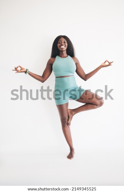 Cheerful African American woman in sportive\
tracksuit practicing balance pose in tree asana during yoga in\
white aerobic studio, happy dark skinned fit girl enjoying body\
positive and vitality