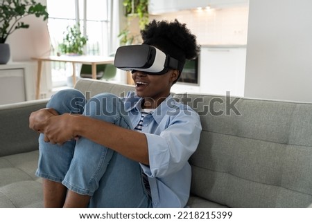 Cheerful African American woman use VR helmet to watch digital movies or visit metaverses in asset space. Impressed girl uses innovative technology to view 3D series sits on sofa in living room