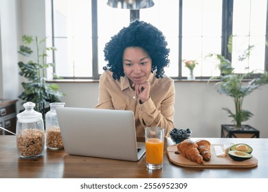 Cheerful African American woman using laptop having breakfast and browsing internet sitting at table in modern kitchen indoors. Successful freelance career concept. - Powered by Shutterstock