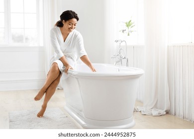 Cheerful african american woman in a white robe sitting on a freestanding bathtub in a neat bathroom - Powered by Shutterstock