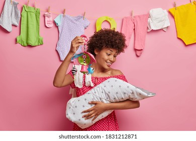 Cheerful African American Woman Young Mommy Cares Of Her Newborn Baby Shows Mobile Toy To Sooth Little Crying Child Poses Against Rope With Washed Infant Clothes. Motherhood And Parenting Concept.