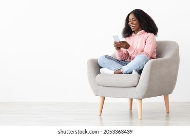 Cheerful african american young lady sitting in arm chair at home, using nice mobile application on brand new smartphone, shopping online, white background, panorama with copy space - Shutterstock ID 2053363439
