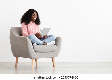 Cheerful african american young lady sitting in arm chair at home, using nice mobile application on brand new digital tablet, shopping online, white background, panorama with copy space