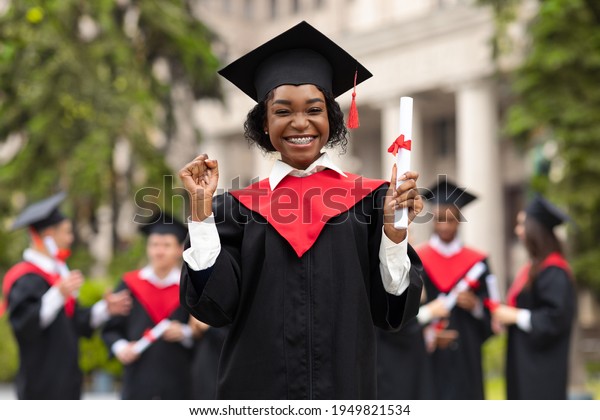 Cheerful african american young lady in graduation\
costume showing her diploma and smiling at camera, black female\
student posing over international group of students at university\
campus, copy space