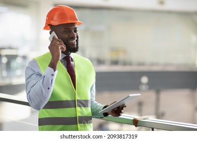 Cheerful african american young male architect in safety workwear having phone conversation with colleagues, using digital tablet, looking at copy space and smiling, side view, panorama