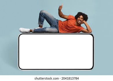 Cheerful african american young man relaxing on huge horizontal brand new cellphone with empty screen and showing thumb up, mockup, black guy enjoying newest entertaining mobile app