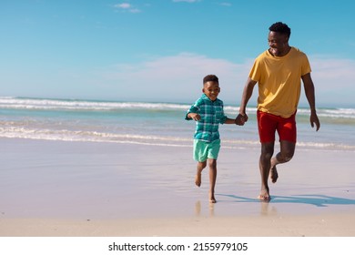Cheerful african american young man and son holding hands while running at beach against blue sky. - Powered by Shutterstock