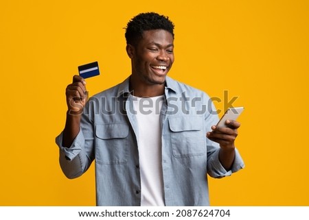 Cheerful african american young man holding brand new cellphone and credit card, trading or gambling online, recommending banking or shopping mobile app, yellow studio background, mockup, copy space