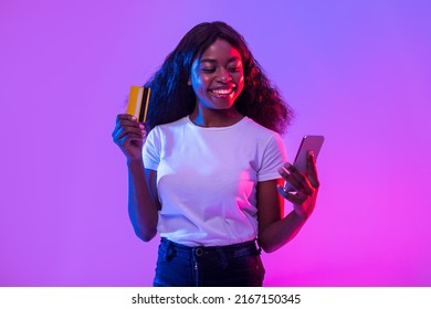 Cheerful African American Young Woman Holding Cellphone And Credit Card, Trading Or Gambling Online, Using Banking Or Shopping Mobile App, Receiving Cashback In Neon Light