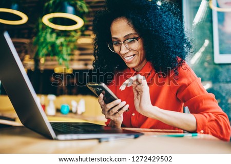 Cheerful African American young woman laughing while reading funny notification on smartphone sitting front laptop computer in cafe. Happy female sending money via online banking app on cellphone