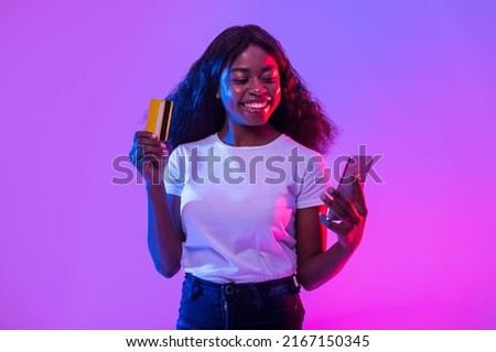 Cheerful African American young woman holding cellphone and credit card, trading or gambling online, using banking or shopping mobile app, receiving cashback in neon light