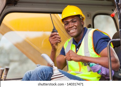 Cheerful African Bulldozer Operator Talking On Walkie Talkie On Construction Site