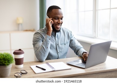 Cheerful African Businessman Talking On Phone Sitting At Laptop Working In Modern Office. Black Guy Having Business Call Conversation By Cellphone At Workplace Concept. Mobile Communication Concept