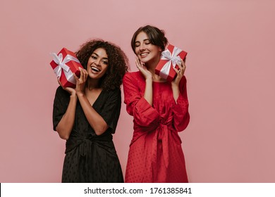 Cheerful african curly girl in polka dot black dress smiling, holding gift box and posing with bright lady in red clothes.