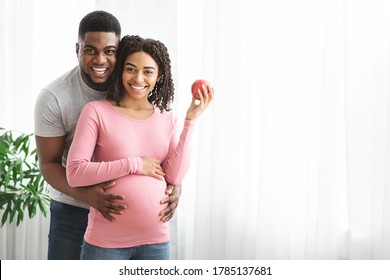 Cheerful african expecting couple posing at home next to window, lady holding fresh apple, copy space