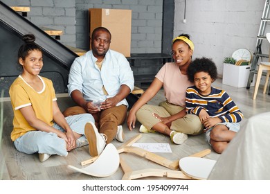 Cheerful African family of father, mother and two children sitting on the floor of large living-room in their new flat and assembling chair - Shutterstock ID 1954099744