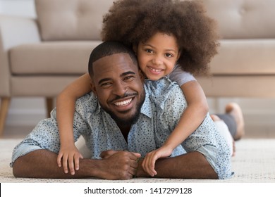 Cheerful african family single black father and cute funny little child daughter portrait, happy dad lying on carpet floor carrying small mixed race kid girl on back smiling looking at camera at home