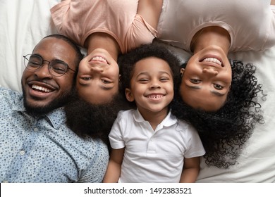 Cheerful african full family married couple with children son and daughter lying down together on bed white bedding smiling looking at camera feels happy. Concept of love, offspring, close up top view