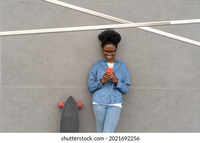 Cheerful African Girl In Trendy Glasses Read Message In Smartphone Or Texting With Friend Using Mobile App And 5g Internet Outdoors. Casual Skateboarder Female Check Social Media After Longboarding