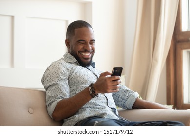 Cheerful African Guy Sitting On Couch In Cozy Living Room Spending Free Time Using Smart Phone Read Joke Message From Friend Laughing Watching Funny Video Online, Enjoy Internet Applications Concept