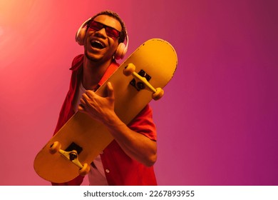Cheerful african guy in sunglasses   headphones posing and skateboard isolated over gradient neon studio background