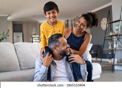 Cheerful african mother and indian father playing with son at home. Cute boy enjoying sitting on father shoulder while looking at camera. Middle eastern family having fun together on the sofa at home.