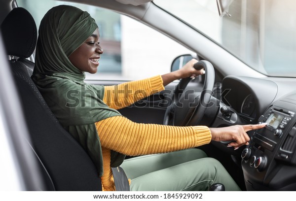 Cheerful African Muslim Woman In Hijab Changing\
Radio Station While Listening Music In Car, Enjoying Automobile\
Audio System, Smiling Islamic Lady In Headscarf Testing Her New\
Vehicle, Side View