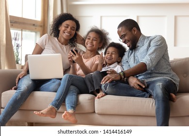 Cheerful African Parents And Kids Laugh Use Devices Together Sit On Sofa, Tech Addicted Family With Children Hold Laptop Phone Digital Tablet Having Fun With Gadgets At Home, Technology Dependence