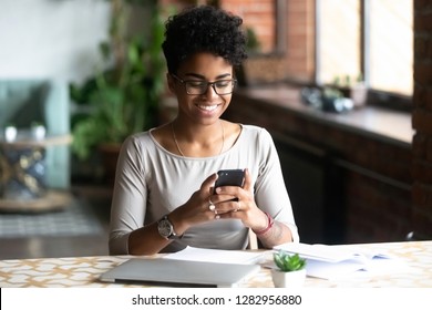 Cheerful african student black woman sitting at table studying using laptop reading a book, take a break holding mobile phone surfing internet received message from friend chatting about weekend plans