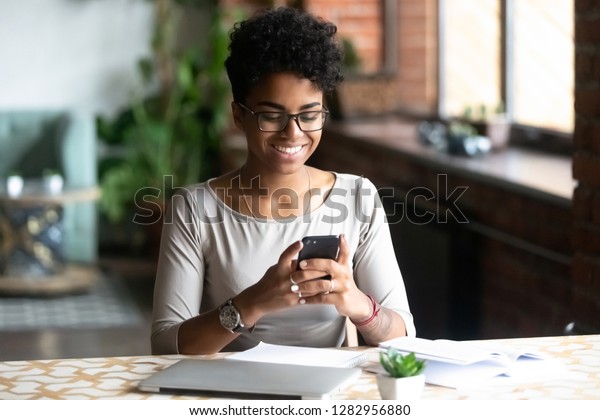 机に座って勉強している明るいアフリカ人学生の黒人女性が ノートパソコンを使って本を読みながら勉強し 休みを取り 携帯サーフィンのインターネットを持つと 週末の計画について友人からの話を受け取った の写真素材 今すぐ編集