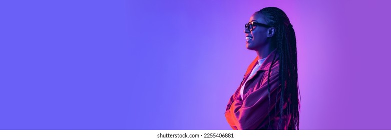 Cheerful african woman in glasses posing  looking away and smile over gradient blue purple background in neon light  Banner  flyer  Concept emotions  facial expression  sales  ad  fashion