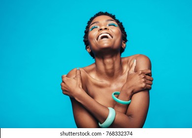 Cheerful african woman hugging herself against blue background. Female model wearing vibrant makeup laughing in studio.