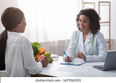 Cheerful African Woman Nutritionist Talking To Female Patient And Taking Notes, Clinic Interior