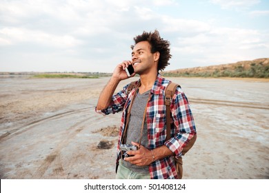 Cheerful African Young Man With Old Vintage Photo Camera Standing And Talking On Cell Phone