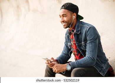 Cheerful african young man wearing cap walking on the beach and chatting by his phone while listening music with earphones.