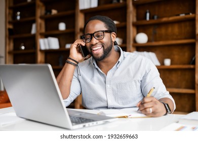 Cheerful African-American Guy Is Doing Multi Tasks Work, A Mixed-race Man Wearing Smart Casual Wear Talking On The Phone, Taking Notes And Using A Laptop On The Workplace In Modern Office