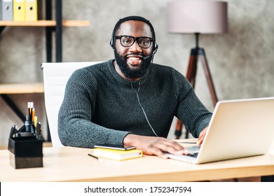 Cheerful African-American Guy Uses A Handsfree Headset And Laptop To Talk Online At His Workplace, Black Confident Man In Glasses Sits At The Office Desk, Looks At Camera With Pleasant Smile