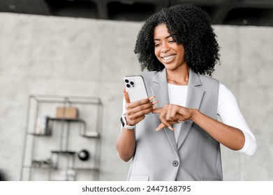 A cheerful African-American woman in a stylish gray blazer looks at her smartphone with a bright smile in a modern office setting, exuding professionalism and approachability. - Powered by Shutterstock