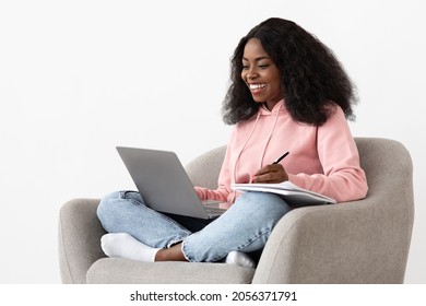 Cheerful African-american Young Woman Attending Webinar From Home, Sitting In Comfy Arm Chair, Using Laptop And Taking Notes, White Background, Copy Space. E-education, Training, Online Course