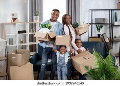 Cheerful Afro American Family Holding Boxes While Standing At Bright Modern Apartment. Happy Family Moving To New House. Concept Of Property And Real Estate.