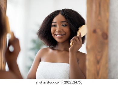 Cheerful Afro Woman Brushing Her Curly Hair With Wooden Brush In Front Of Mirror At Home. Young Black Lady In Bath Towel Making Morning Beauty Routines, Taking Care Of Herself Indoors