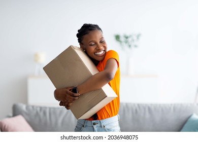Cheerful Afro woman hugging carton parcel, receiving long awaited delivery, getting online order indoors. Satisfied female customer empracing her internet purchase in cardboard package - Shutterstock ID 2036948075