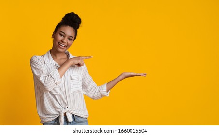 Cheerful Afro Woman Pointing At Something On Her Open Palm, Demonstrating Invisible Object, Free Place For Mockup, Yellow Background, Panorama