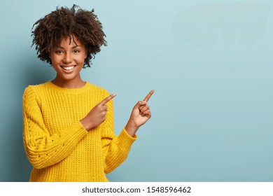 Cheerful Afro woman points away on copy space, discusses amazing promo, gives way or direction, wears yellow warm sweater, has pleasant smile, feels optimistic, isolated over blue background - Shutterstock ID 1548596462